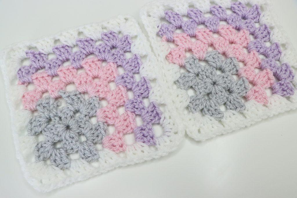 A pair of mitered granny squares in pastel shades of pink, purple and grey sit on a white surface.