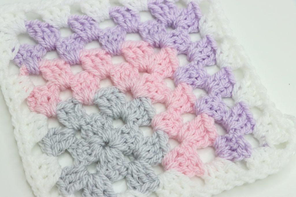 Close up of a mitered corner granny square with cream, purple, pink and grey yarn.