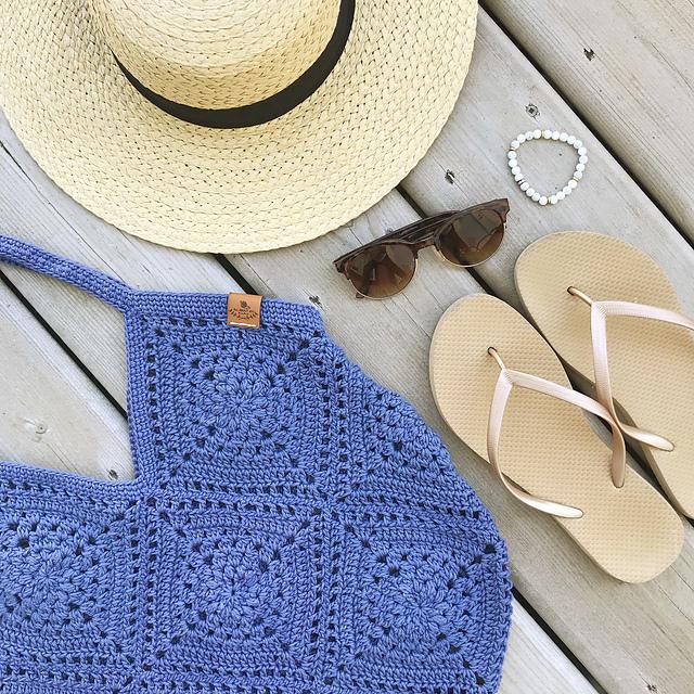 A blue crochet bag sits on a wooden background alongside a pair of flip flops, sunglasses and a sunhat.