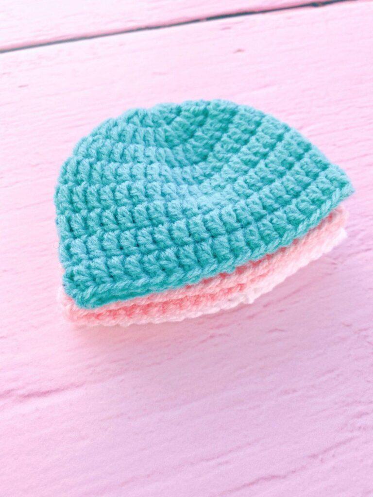A small stack of child-sized beanie hats sits on a pink background. They are made from pastel-coloured yarn.