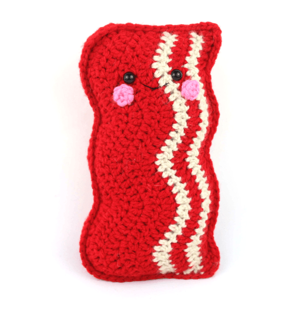 A slice of crocheted bacon is displayed against a white background. The bacon has a cute face and rosy cheeks. 