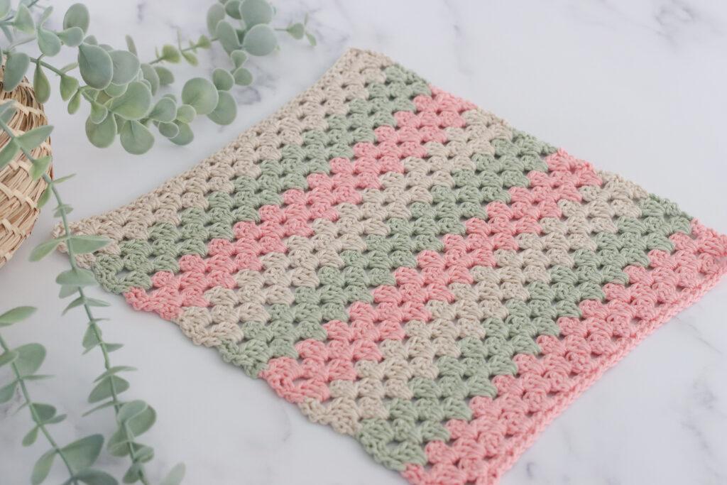 a crochet swatch of the granny stripe stitch in three colours, pastel green, pink and cream on a marbled backdrop and foliage to the left of the image. 