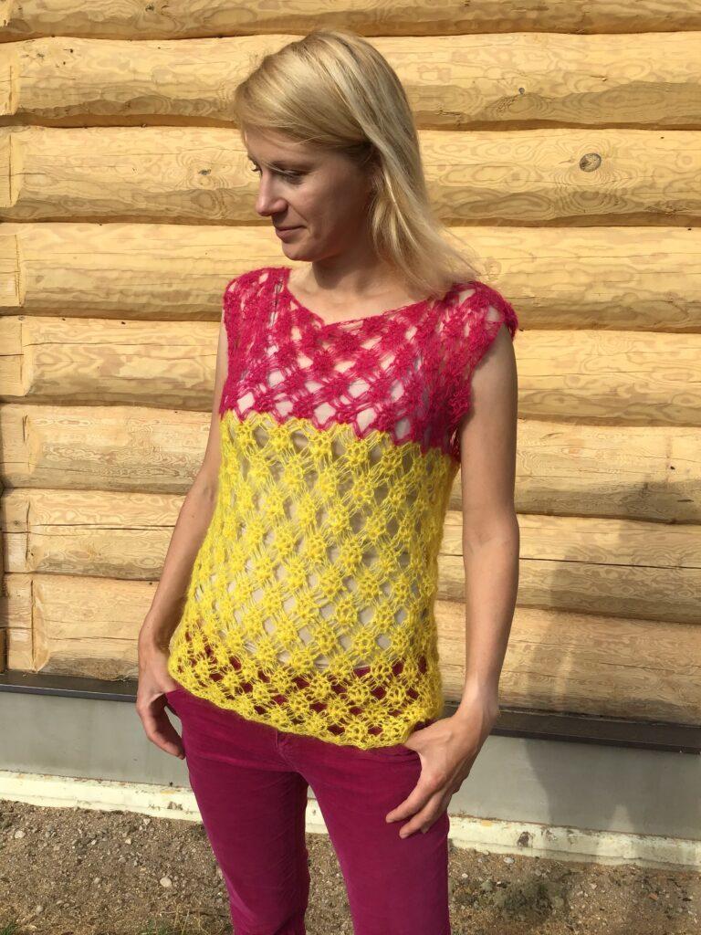 A woman stands in front of a log cabin wearing red jeans and a crocheted yellow and pink tee with no sleeves.