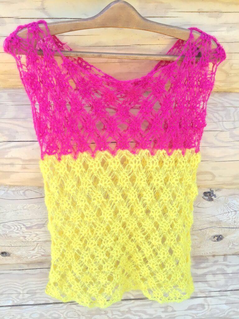 A crocheted yellow and pink tee with no sleeves is hanging on a log cabin wall.