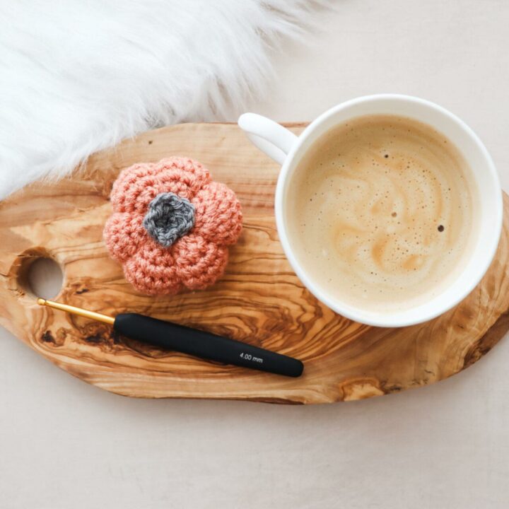 a flat lay image of a crochet pumpkin made in burnt orange yarn lay next to a cup of coffee on a wooden slice with a black and gold crochet hook