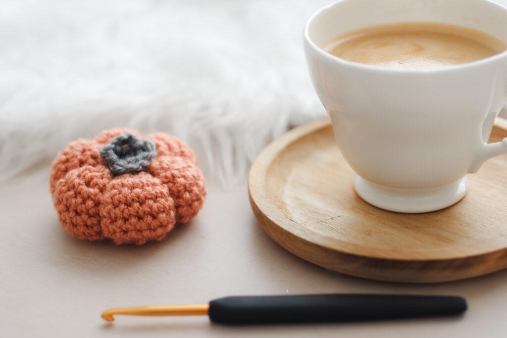 a crochet pumpkin made in burnt orange wool lay next to a cup of coffee on a wooden coaster with a black and gold crochet hook in the foreground lay horizontal
