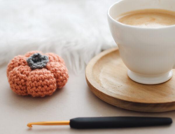 a crochet pumpkin made in burnt orange wool lay next to a cup of coffee on a wooden coaster with a black and gold crochet hook in the foreground lay horizontal