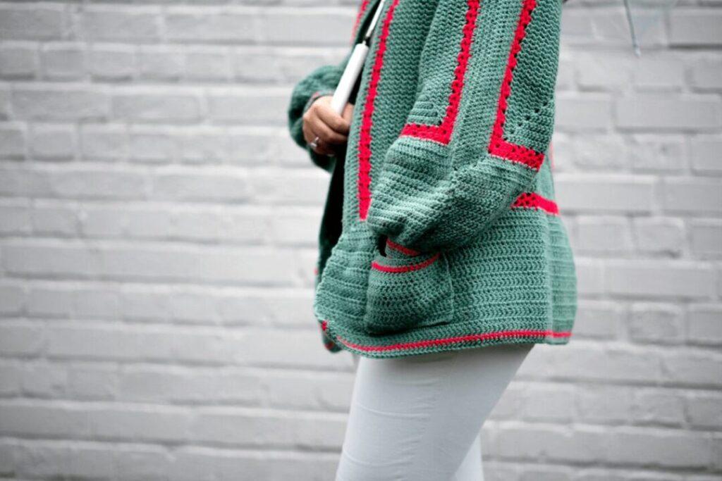 A woman wears a green and red cardigan made from a giant crochet granny square.