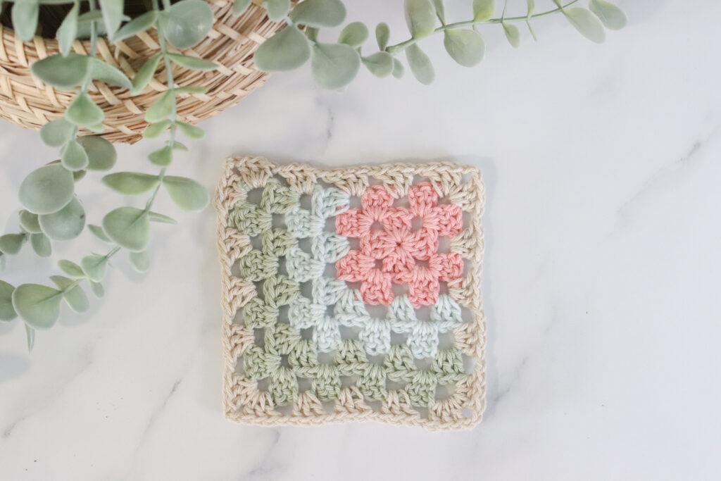 A mitred granny square in pink, blue, green and oatmeal coloured yarn on a marble background with plants surrounding it.