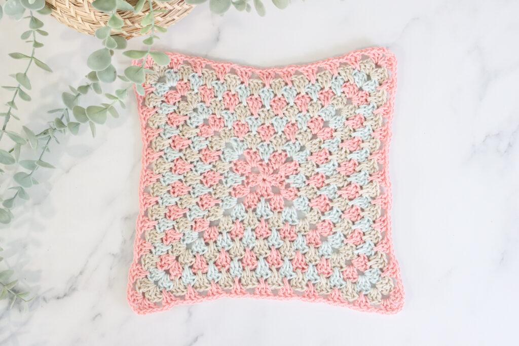 flat lay image of a spiked crochet granny square on a marble backdrop with foliage in the top left corner. The spiked granny square is made of three alternating colours, pale pink, pale blue and beige.