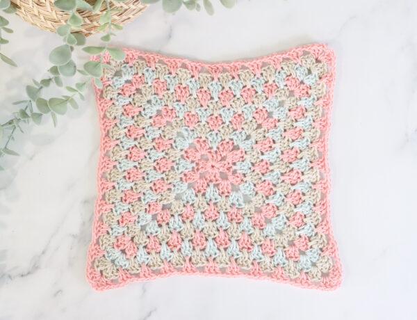 flat lay image of a spiked crochet granny square on a marble backdrop with foliage in the top left corner. The spiked granny square is made of three alternating colours, pale pink, pale blue and beige.