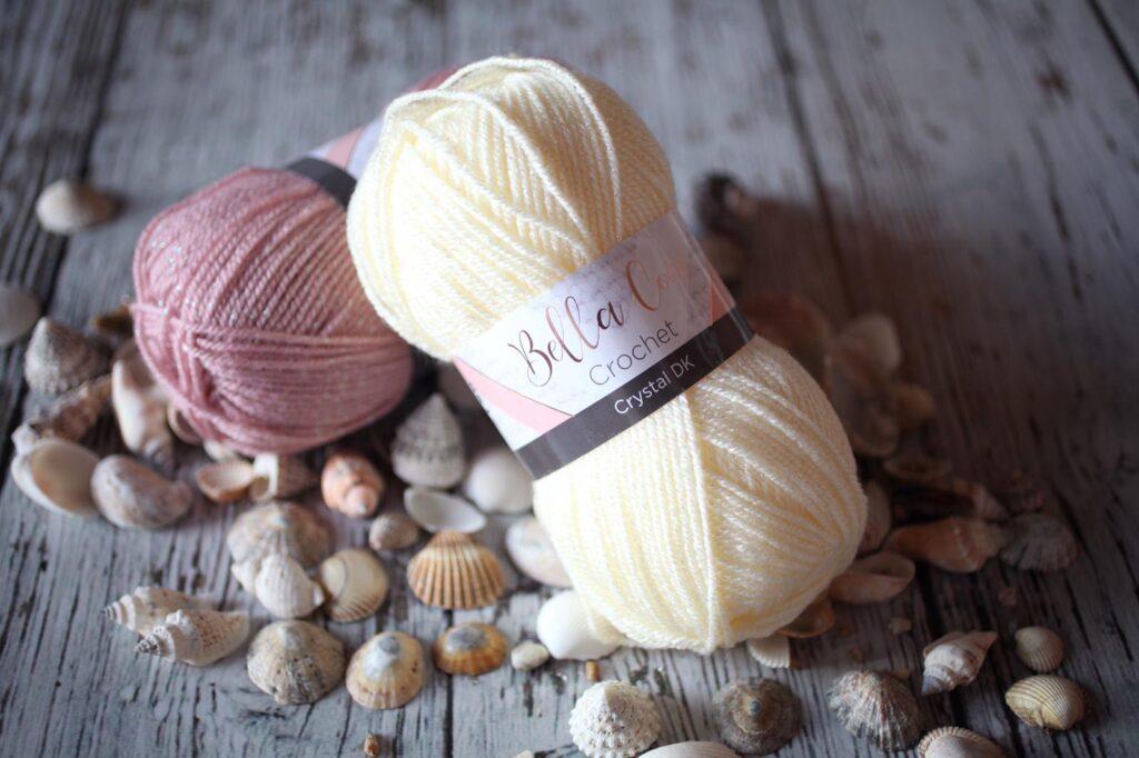 A pair of DK yarn balls sit on top of shells. The yarns are cream and pink coloured with sparkles.