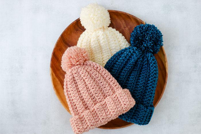 A stack of crochet hats with pompoms sits in a wooden bowl. They are neutral shades and made from thick wool.