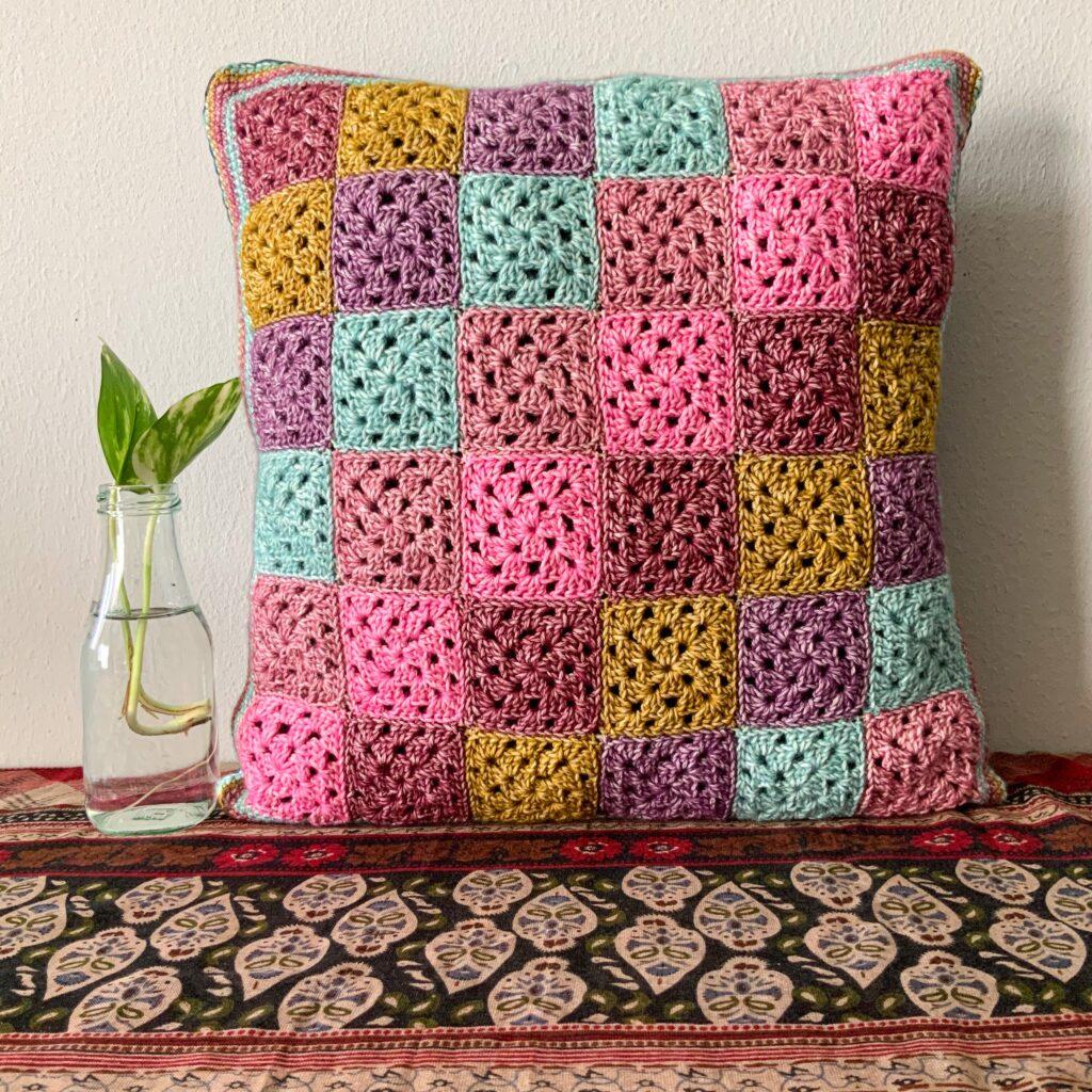 A colourful cushion is made from mini granny squares. It sits on a surface beside a small plant sprout.