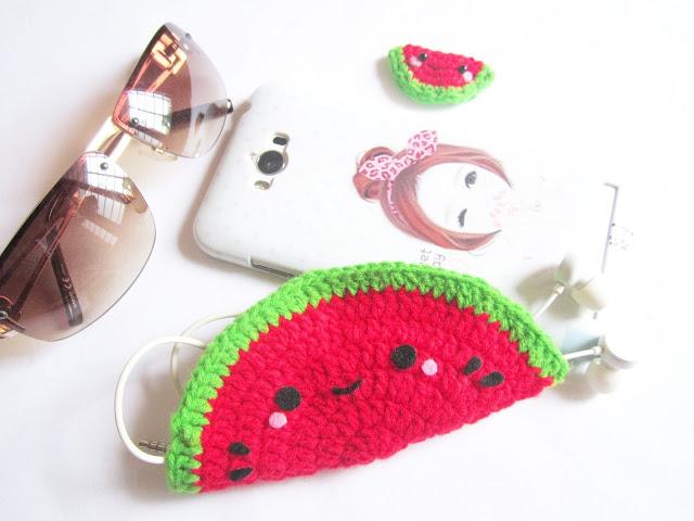 A crochet pouch shaped like a watermelon sits on a white surface beside a phone and sunglasses. The pouch contains headphones.