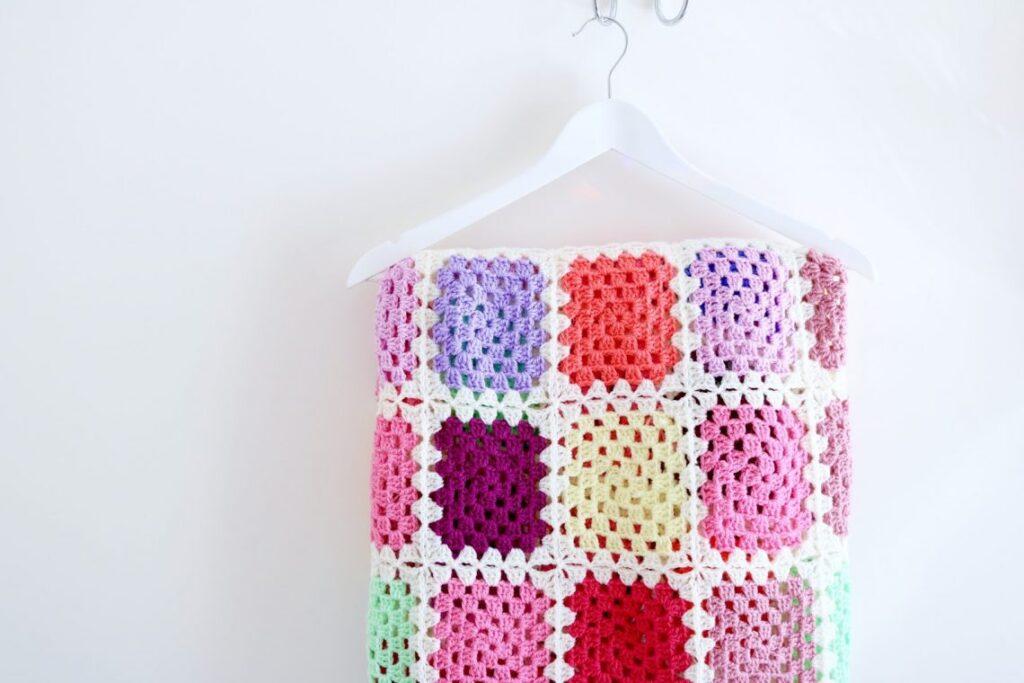 A granny square blanket in lots of pastel tones is hanging on a white wall.