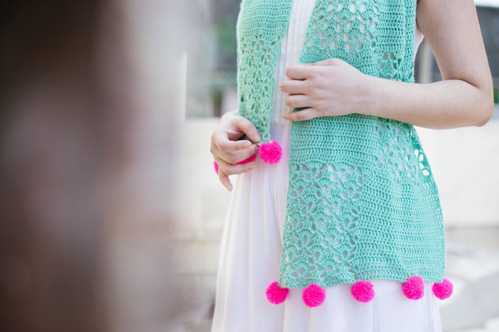 A woman is wearing a white dress and a seafoam-coloured crochet shawl with panels of lace and hot pink pompoms on the end. 