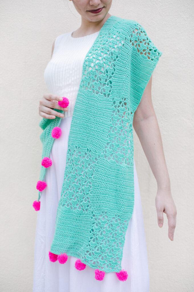 A woman is wearing a white dress and a seafoam-coloured crochet shawl with panels of lace and hot pink pompoms on the end. 