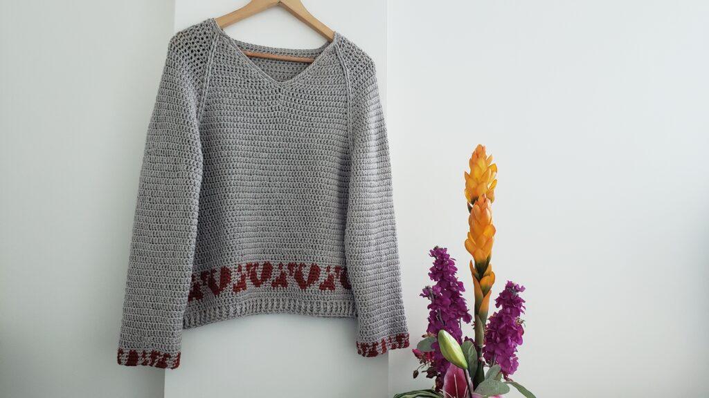 A taupe crochet sweater with a deep red animal trim motif on the bottom hangs up against a white wall beside a bouquet of tropical flowers in shades of orange and pink.