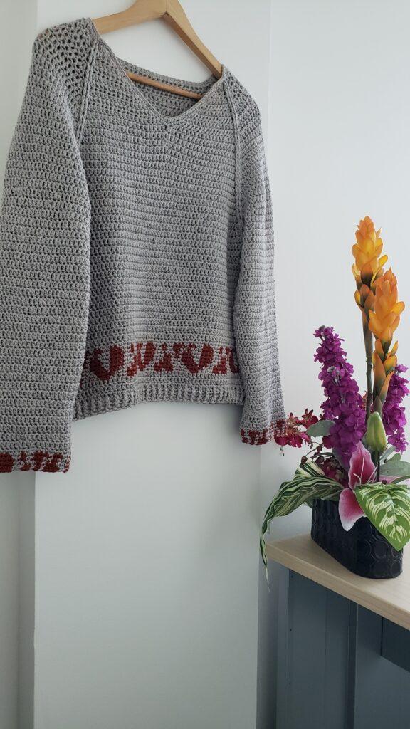 A taupe crochet sweater with a deep red animal trim motif on the bottom hangs up against a white wall beside a bouquet of tropical flowers in shades of orange and pink.