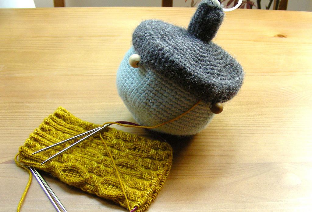 A project bag is fashioned in the shape of an acorn. The bag is made from crochet and holds a knitted sock.