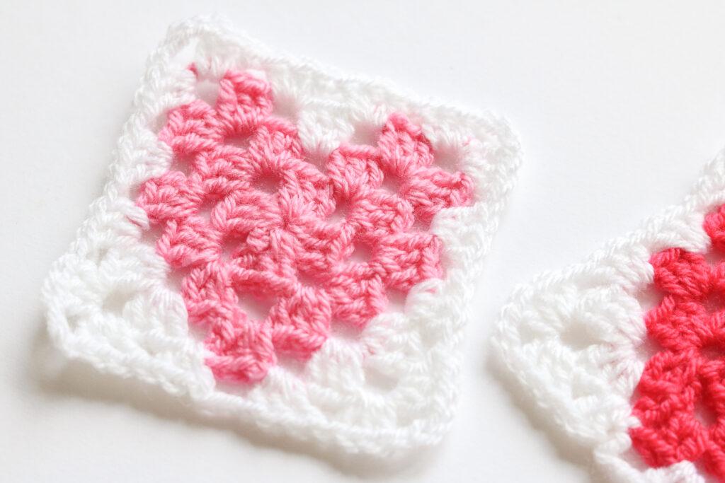 A pair of crochet granny squares with central heart motifs sit on a white background. 