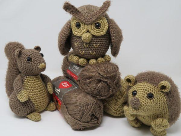 A trio of Autumn-themed crochet animals sit on a stack of brown-toned yarn. There is a squirrel, an owl and a hedgehog. 
