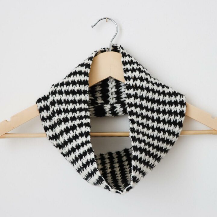 a wooden hanger on a white wall with a black and white houndstooth crochet cowl around the neck of the hanger.