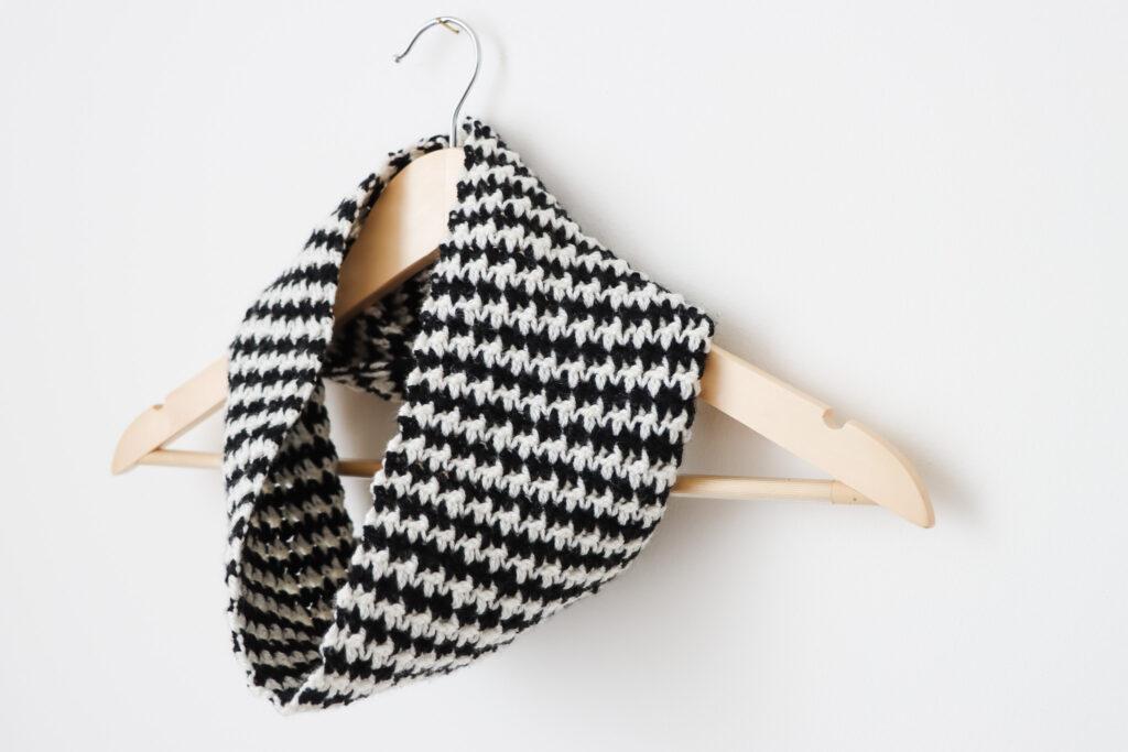 a wooden hanger on a white wall with a black and white houndstooth crochet cowl around the neck of the hanger. 