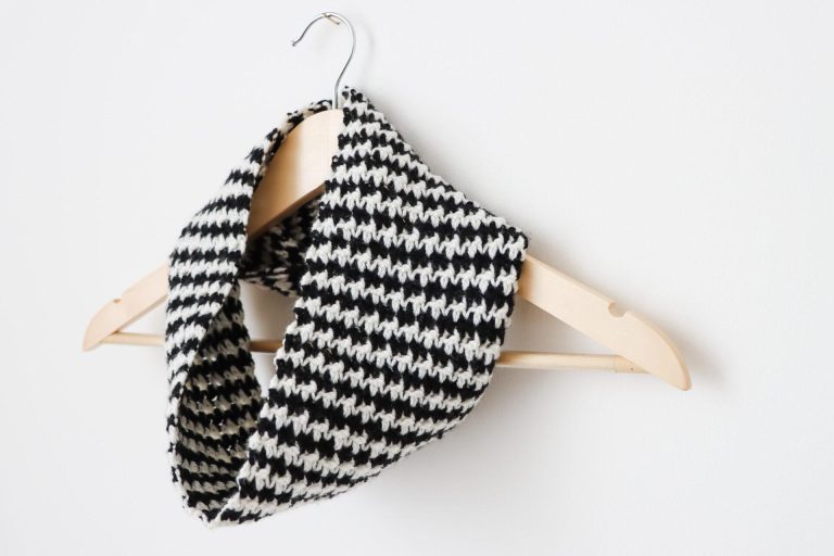 a wooden hanger on a white wall with a black and white houndstooth crochet cowl around the neck of the hanger.