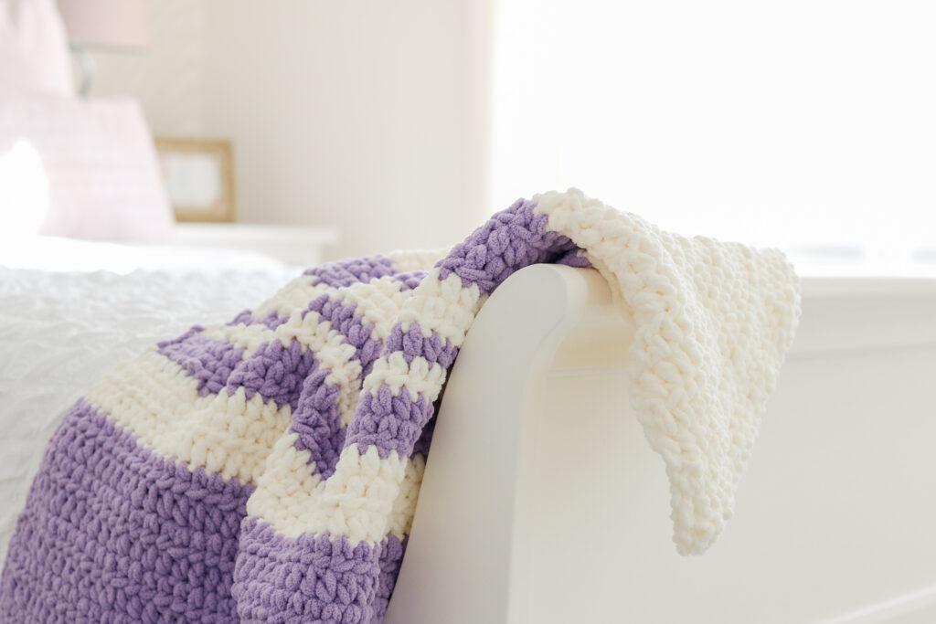 A lilac and white blanket made from crochet stitches is draped over a white bed. 