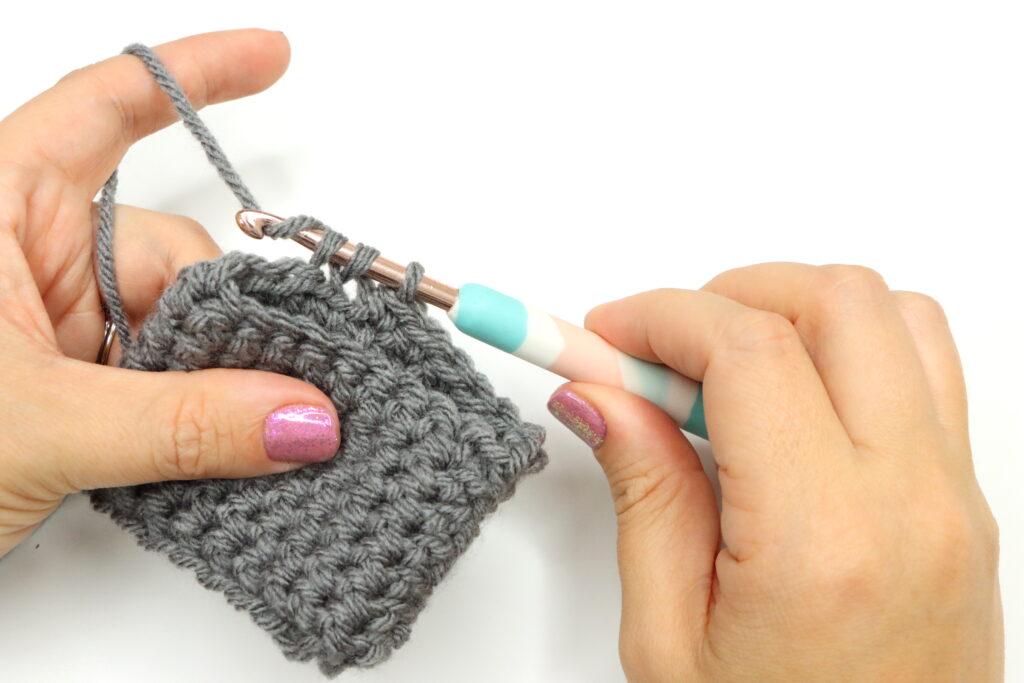 A woman holds a grey swatch of crochet and a pastel-coloured hook. She is working into the stitches.