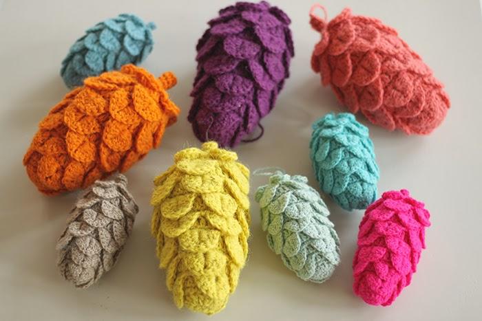 A display of multi-coloured crochet pinecones sit on a white surface. They are made from jewel tones of green, blue, pink, purple, green and orange.