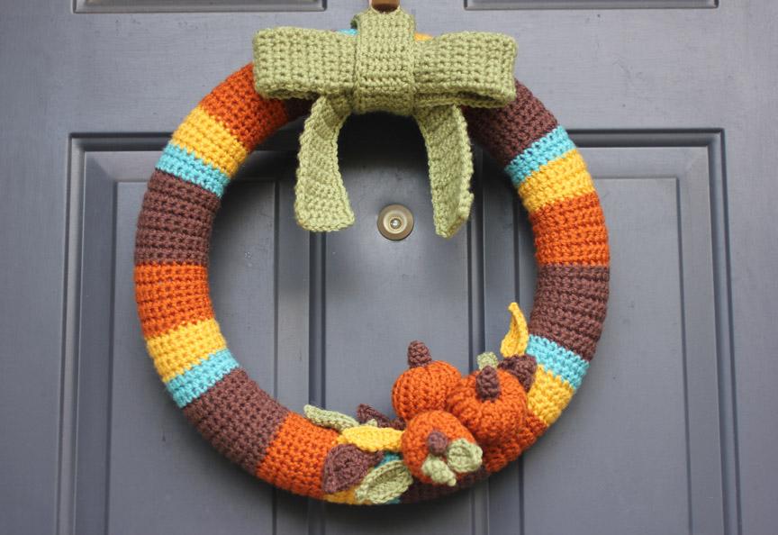 A crochet wreath in Fall colours of brown, orange, yellow, blue and green sits against a black door. There are small crochet pumpkins on the bottom of the wreath.
