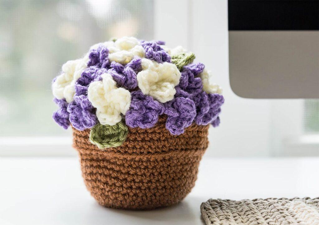 A crocheted pot of purple and white flowers sits on a white surface.