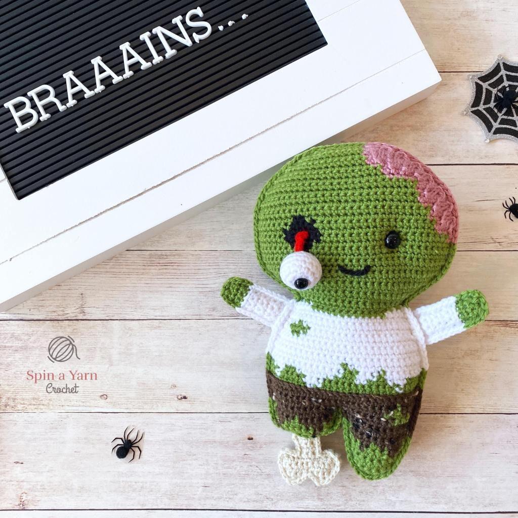 A green crochet zombie with a loose eyeball and visible brains sits on a wooden background beside a black sign with white writing which reads, " Braaains"