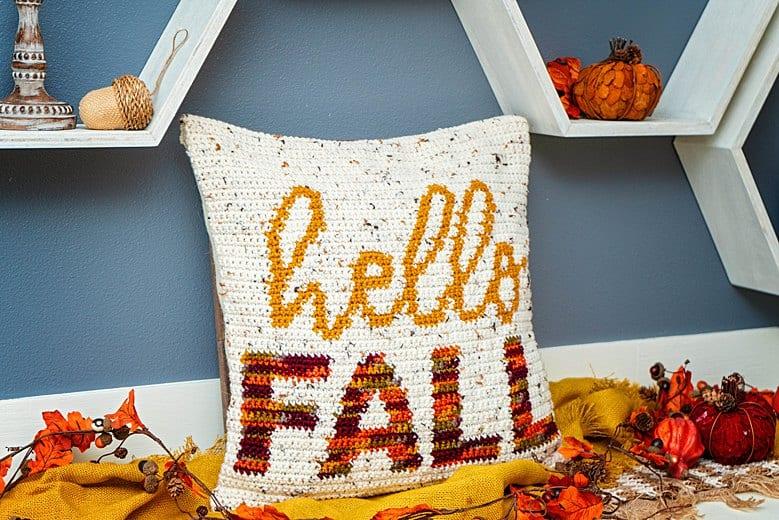 A cream crochet cushion with the words Hello Fall on sits against a blue wall surrounded by autumn foliage.