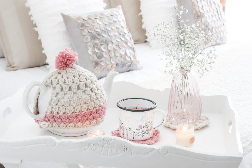 Decorative tea cosy with mug, candle, cushions and a vase of flowers.