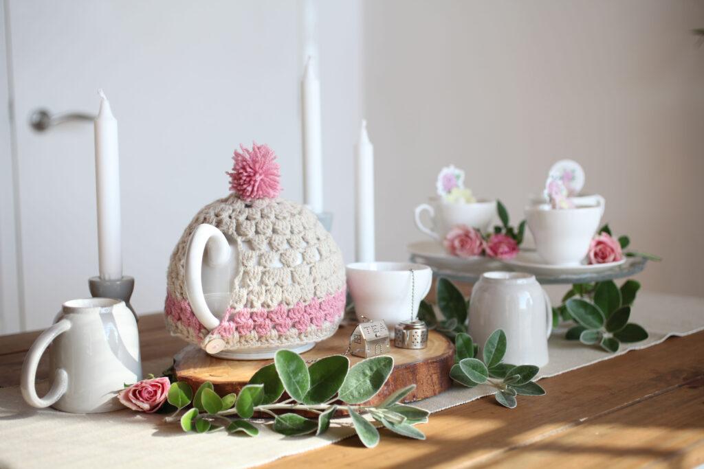A crochet tea cosy sits on a teapot surrounded by cups and saucers