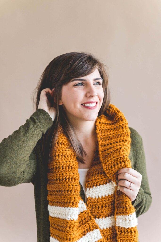 a woman in a green cardigan wears a yellow and white crochet scarf. It is made in chunky yarn and looks cosy.