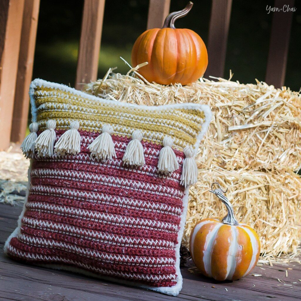 A tasselled cushion sits against a hay bale, surrounded by pumpkins.