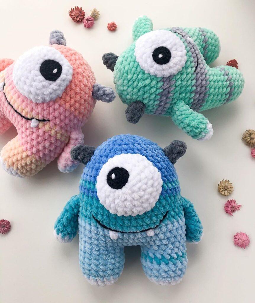 A trio of one-eyed monsters sit in a circle. They are crocheted from velvet yarn in pastel colours.