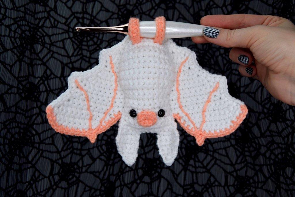 A pink and peach coloured crochet bat hangs upside down from a white crochet hook.