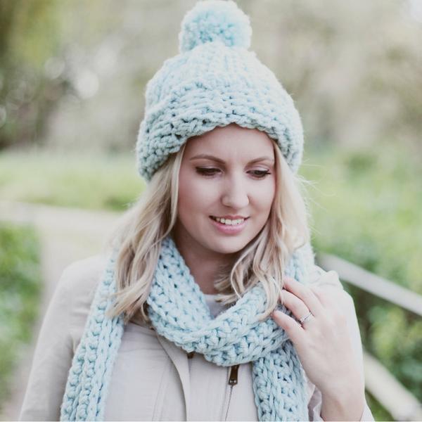 A blonde woman stands outside wearing a blue scarf and hat set. They are made using a matching crochet stitch.