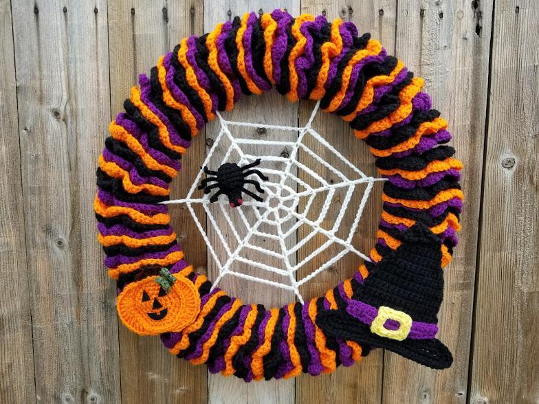 A Halloween-themed wreath made from orange, purple and black yarn is hanging on a wall. It has spider, pumpkin and a witches hat motifs decorating it.