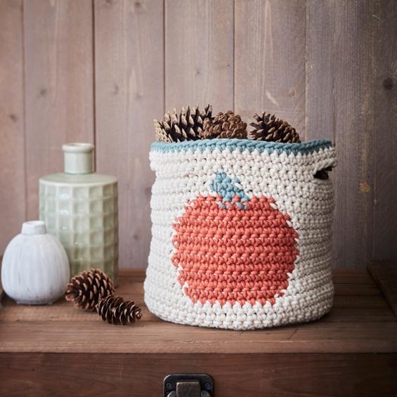 A cream crochet basket with an orange pumpkin motif sits on a wooden shelf. It is filled with pinecones.