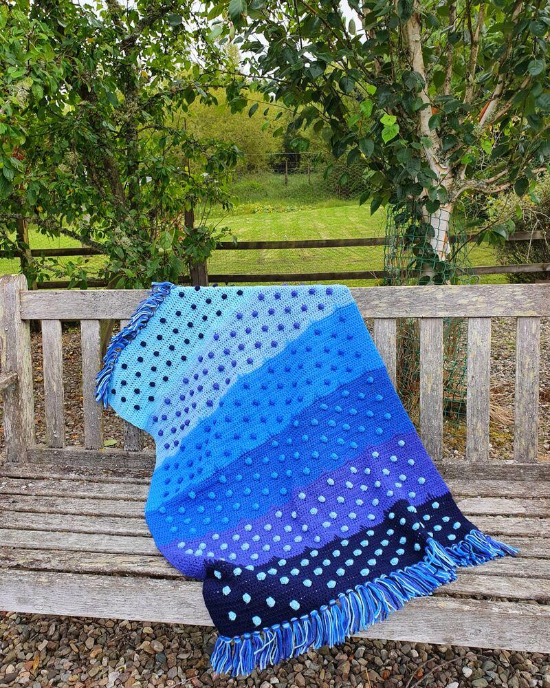 A blue ombre crochet blanket with bobbles is draped over a garden bench outside.