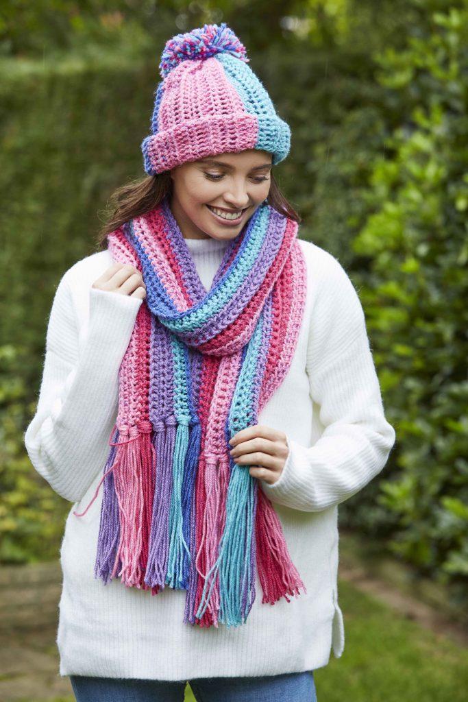 A woman is wrapped up warm in a white sweater and a colourful crochet hat and scarf in shades of purple, pink and blue. 