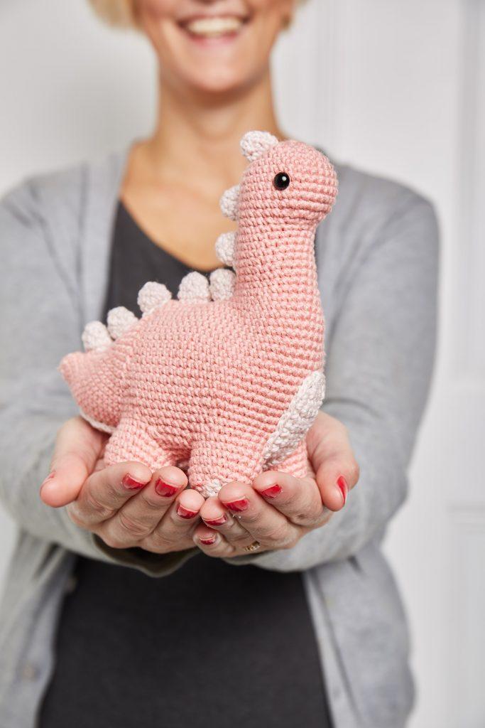A pink coloured crochet dinosaur sits in a woman's hands. He has paler pink spines running along his back.