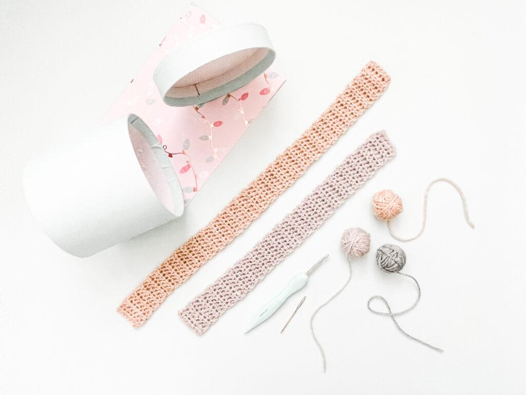 a flat lay image showing two different lengths of crochet ribbon in gold and silver yarn lay flat on a pale blue background with a gift box, pink gift wrap and balls of yarn lying alongside the ribbon 